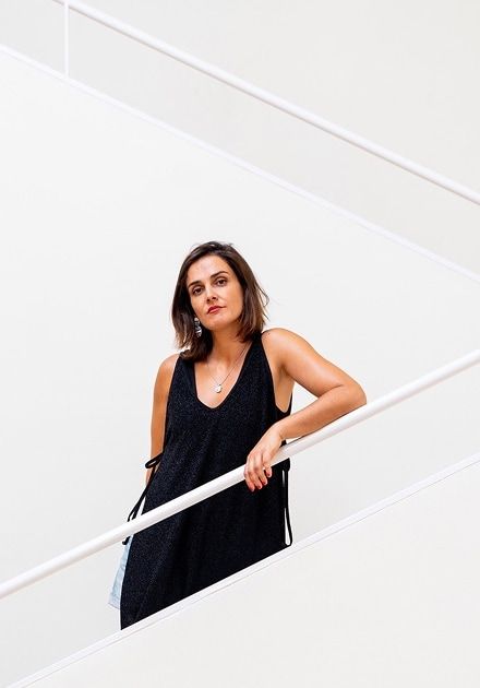 Mulher de cabelo castanho curto, com vestido preto sem mangas e decote em V, encostada a um corrimão branco numa escadaria minimalista de paredes claras, olhando diretamente para a câmara com expressão neutra.