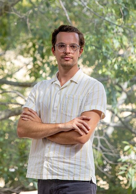 Homem jovem de pé ao ar livre, de braços cruzados, a olhar diretamente para a câmara, usando óculos transparentes, camisa de manga curta às riscas claras e calças escuras, com árvores verdes desfocadas ao fundo.