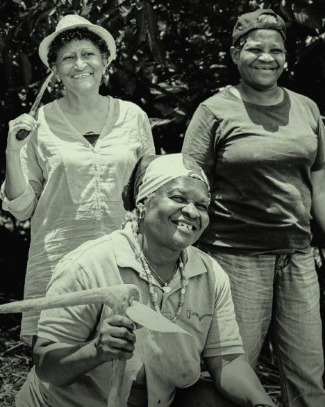 Fotografia a preto e branco de três mulheres trabalhadoras rurais a sorrir para a câmara, ao ar livre. A mulher em primeiro plano está agachada, usa lenço na cabeça, camisa de manga curta e segura uma enxada. Atrás dela, à esquerda, uma mulher de chapéu de palha e camisa larga segura uma ferramenta agrícola ao ombro. À direita, outra mulher de boné e t-shirt escura está de pé com as mãos atrás das costas. Ao fundo vêem‑se árvores e vegetação densa.